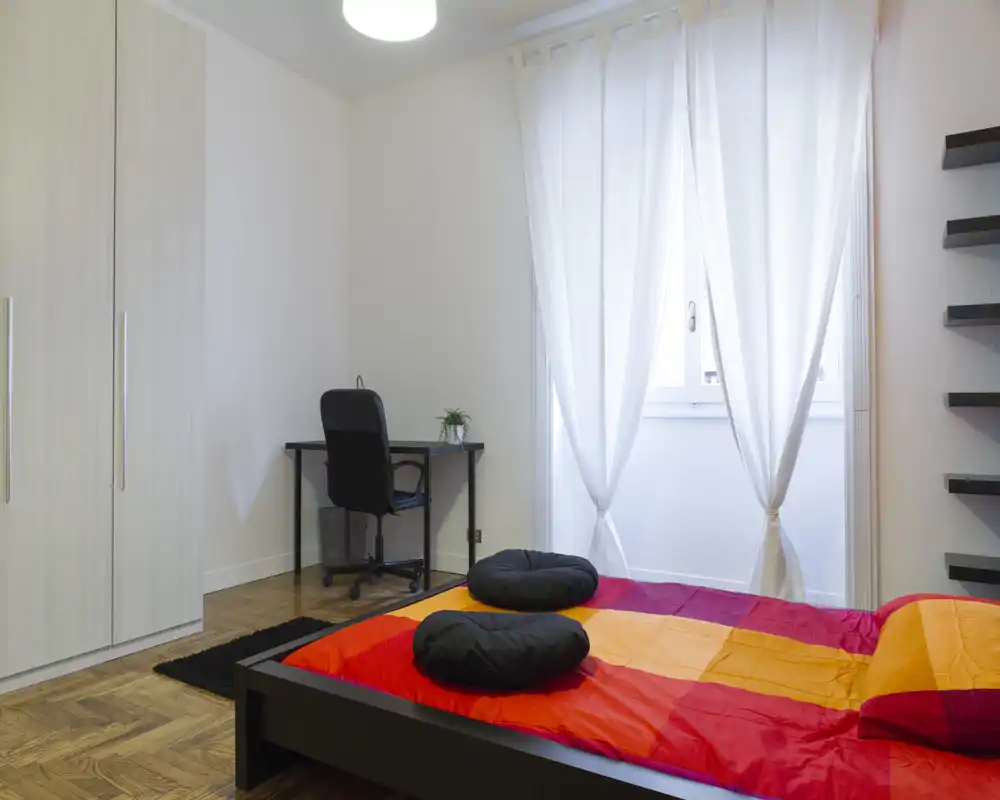 Dormitorio moderno y luminoso con cama baja y ropa de cama colorida, armario, escritorio y luz natural por la ventana con cortinas translúcidas — acogedor y limpio.
