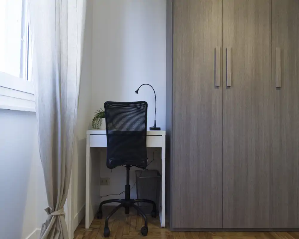 Rincón de dormitorio con escritorio compacto, silla giratoria y gran armario; luz natural y decoración neutra que crean un espacio tranquilo.