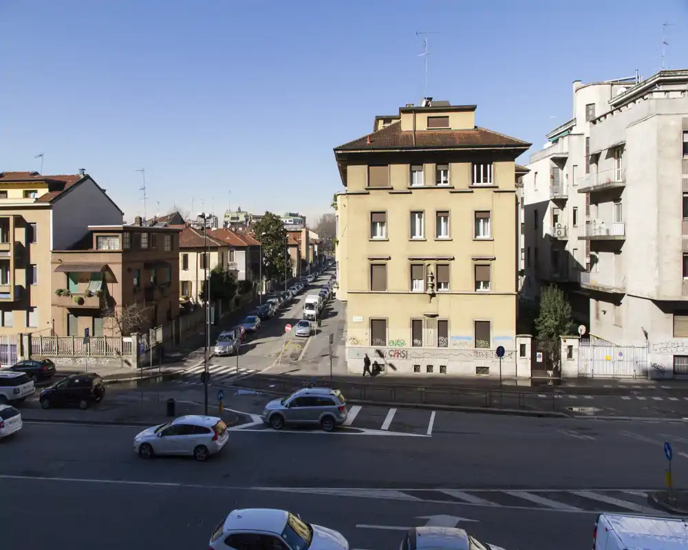 Vista exterior a la calle que muestra una manzana urbana con coches aparcados, edificios de viviendas y cielo despejado — apta para mostrar la vista desde una ventana o balcón.
