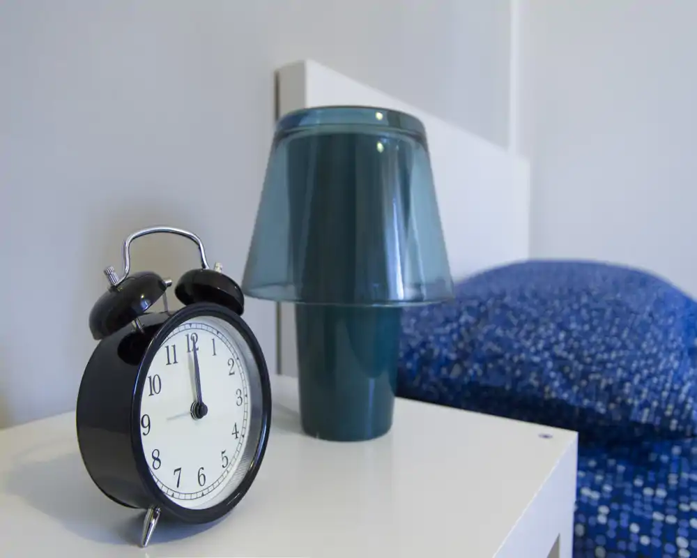 Detalle luminoso de dormitorio con mesita de noche, despertador, una lámpara de sobremesa turquesa y parte de la cama con funda azul — presentación moderna y ordenada ideal para anuncios.