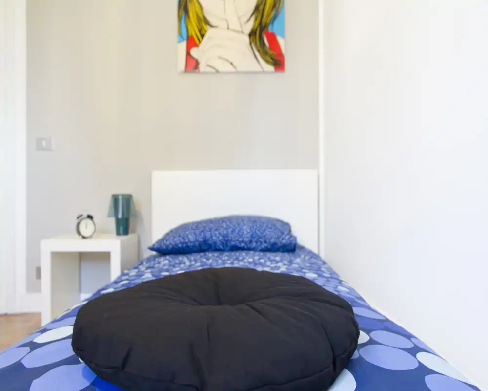Habitación individual ordenada con funda de cama azul estampada, cojín redondo decorativo y mesita minimalista; espacio luminoso, sencillo y acogedor, ideal para un huésped.