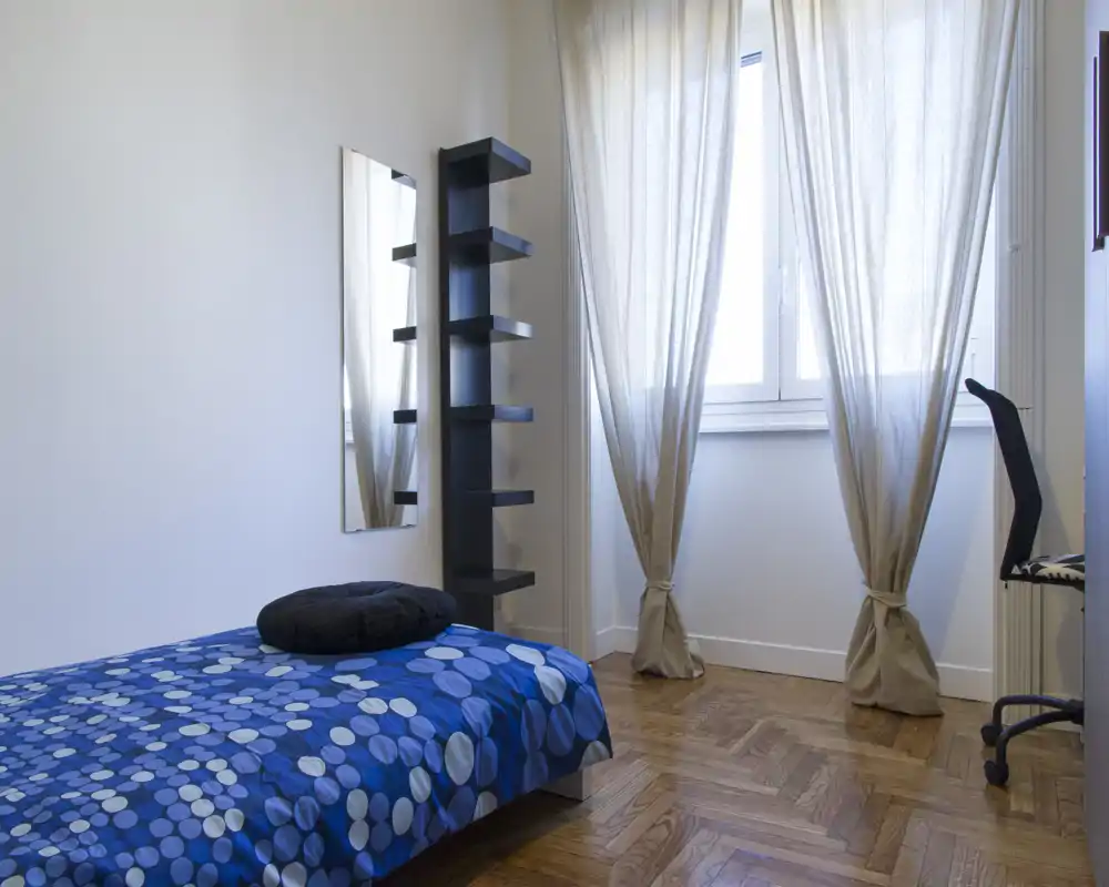 Dormitorio individual sencillo y luminoso con ropa de cama azul estampada, luz natural procedente de una ventana grande y mobiliario moderno y minimalista, incluyendo estantería alta y silla de escritorio.