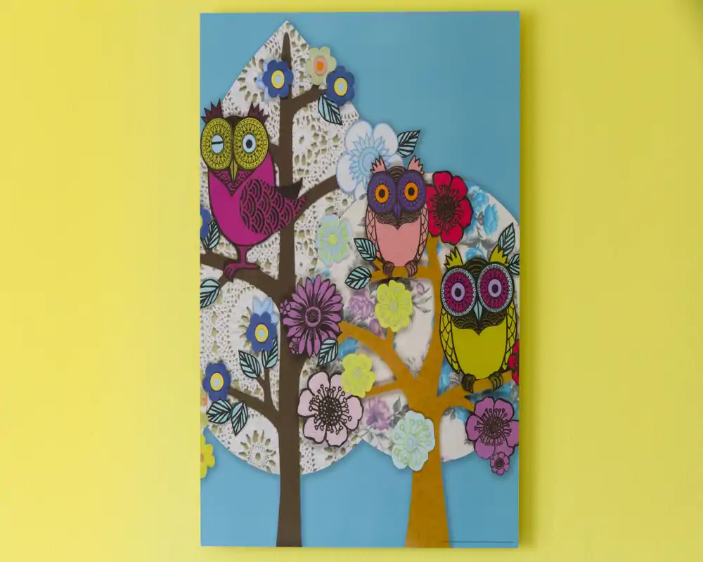 Panel decorativo colorido con búhos estilizados y flores sobre fondo azul, colgado en una pared amarilla.
