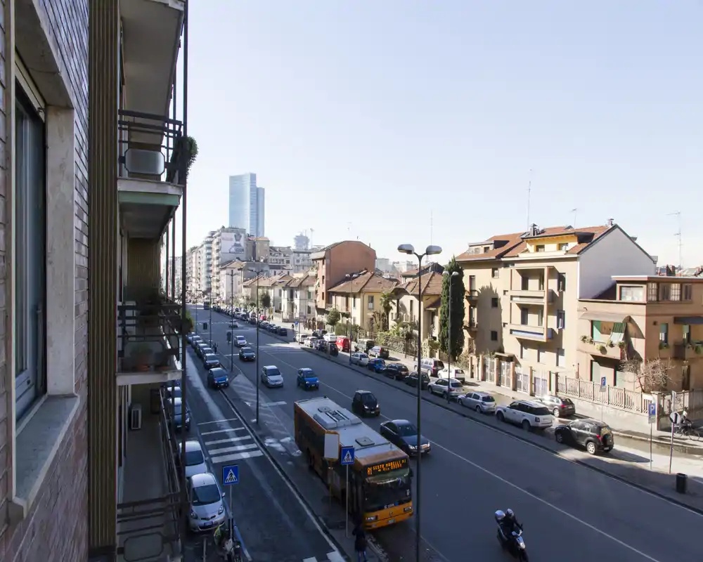 Vista desde el balcón a una calle urbana concurrida con coches aparcados, un autobús y edificios residenciales — escena diurna luminosa, ideal para mostrar la ubicación y el acceso.
