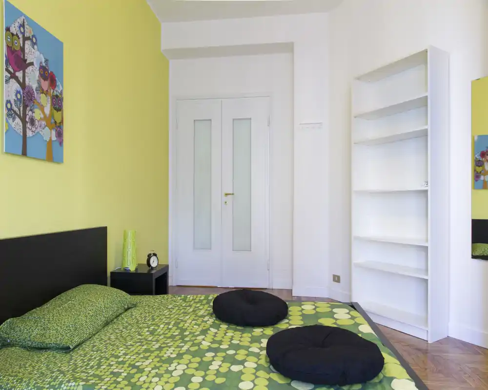 Dormitorio moderno y luminoso con una cama baja grande con ropa de cama verde estampada, cojines redondos decorativos y una mesita minimalista; el espacio incluye un cuadro a juego y una estantería alta vacía.