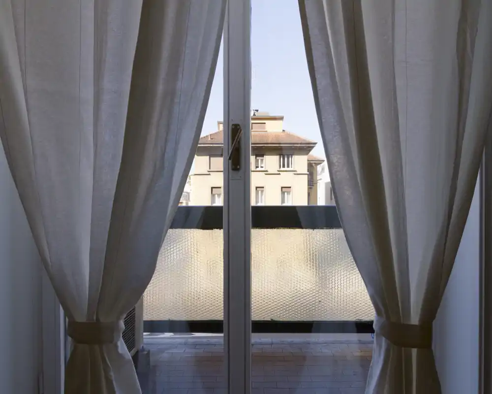 Vista a través de puertas de cristal hacia un pequeño balcón, con cortinas que filtran la luz y edificios vecinos visibles al fondo.