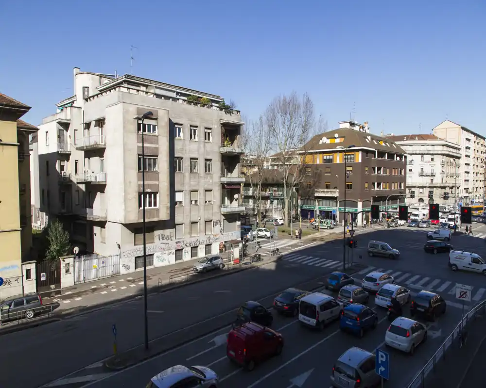 Vista exterior a la calle que muestra una intersección urbana con coches estacionados y en movimiento, edificios residenciales de varias plantas y cielo despejado — útil para ilustrar la ubicación y los alrededores.