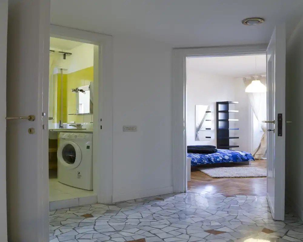 Vista directa del dormitorio desde el pasillo: cama baja con ropa de cama azul, alfombra y estantería; la luz natural ofrece un ambiente agradable.