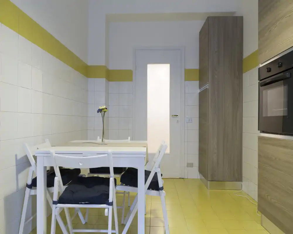 Rincón de cocina luminoso y moderno con mesa blanca y cuatro sillas, muebles efecto madera y horno empotrado; suelo de baldosas amarillas y un jarrón sencillo que aportan una sensación fresca y minimalista.