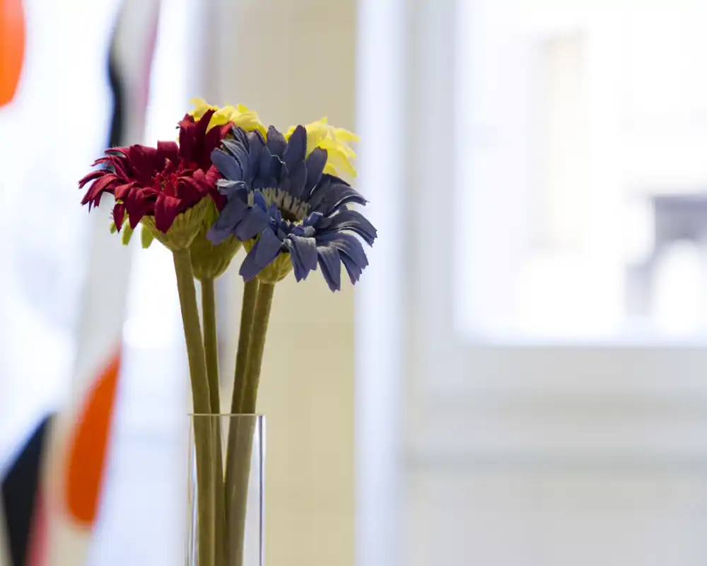 Plano cercano de flores decorativas artificiales en un jarrón transparente colocadas en el interior; el fondo está desenfocado y muestra una ventana luminosa y formas abstractas. La foto se centra en el arreglo floral más que en la función de la habitación.