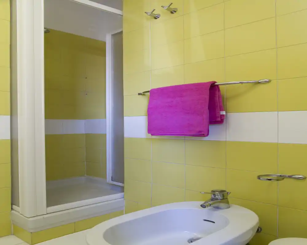 Baño moderno y luminoso con azulejos amarillos, bidé y plato de ducha; una toalla magenta aporta un acento vivo.
