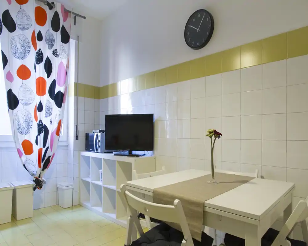 Área de comedor luminosa y ordenada con mesa y sillas blancas, un camino de mesa y un jarrón con una flor; grandes cortinas estampadas y un mueble con TV aportan personalidad.