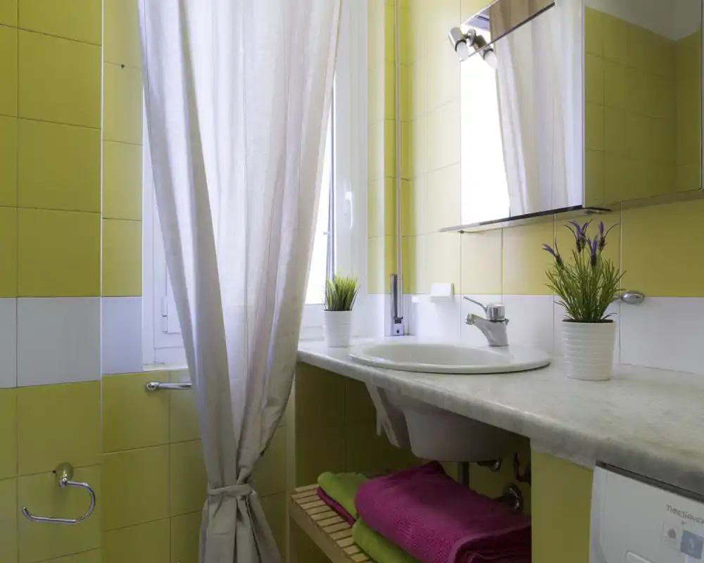 Baño compacto y luminoso con azulejos amarillos, lavabo blanco sobre encimera de mármol y armario espejo. Toallas y pequeñas plantas en maceta ofrecen un aspecto limpio y acogedor.
