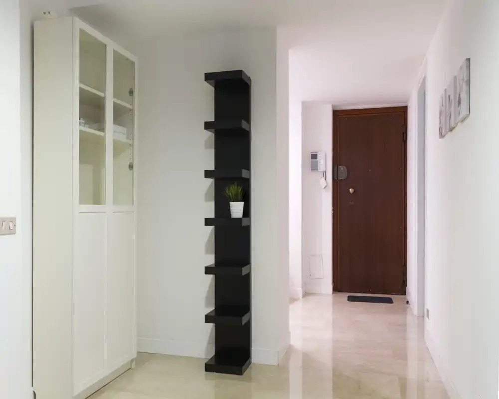 Entrada de apartamento luminosa y minimalista con puerta principal de madera, suelo de baldosas y una estrecha estantería negra junto a un armario blanco.