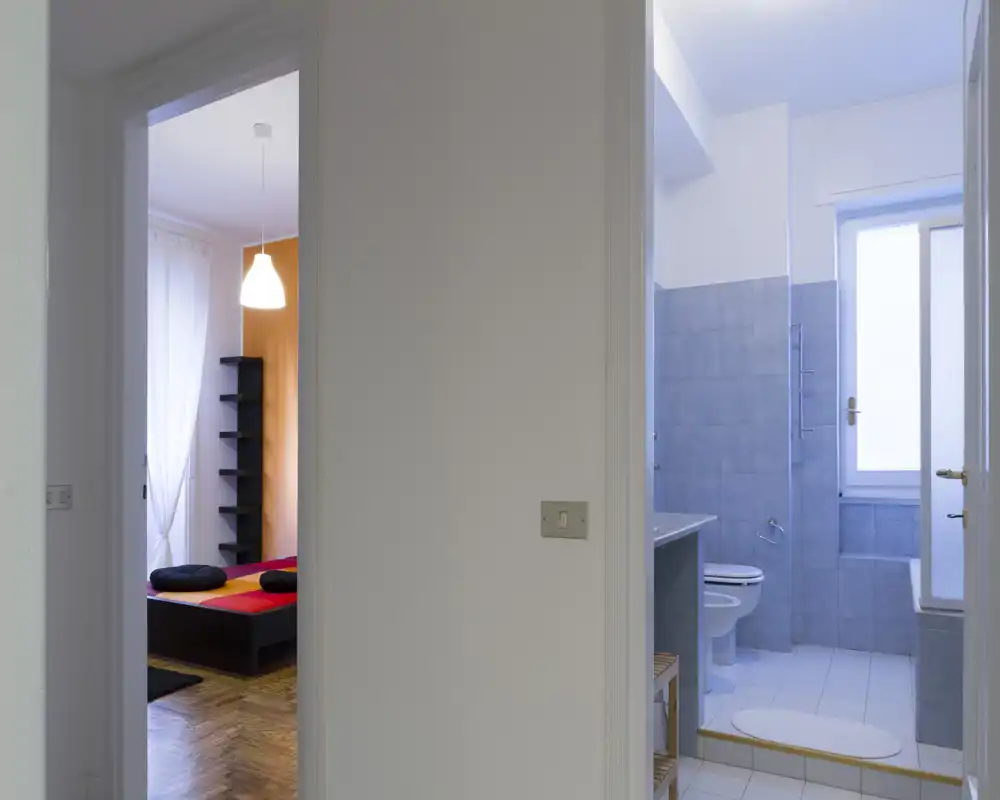 Dormitorio luminoso visto a través de una puerta, con una cama baja con ropa de cama colorida, estantería vertical y suave luz natural de la ventana.