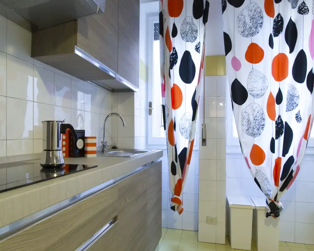 Cocina moderna y compacta con placa integrada, fregadero de acero y cortinas estampadas y coloridas; luminosa y bien presentada para un anuncio.