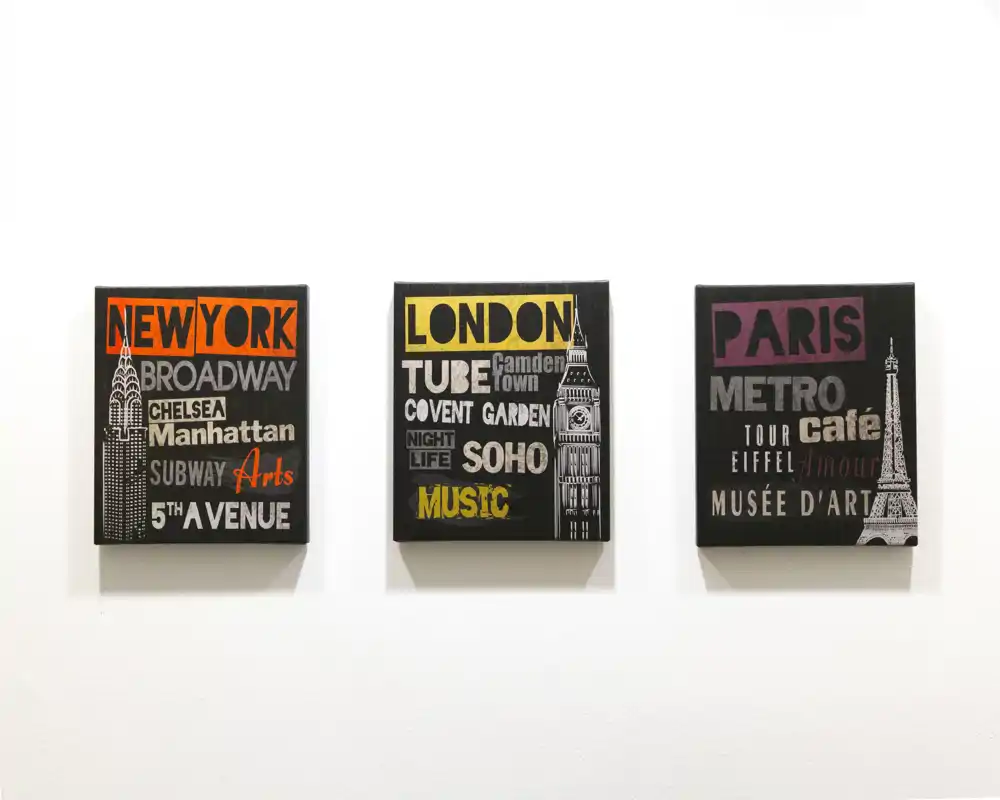 Primer plano de tres lienzos decorativos con temática de ciudades (Nueva York, Londres, París) colgados en una pared blanca; la foto se centra en el arte mural y no en una habitación concreta.