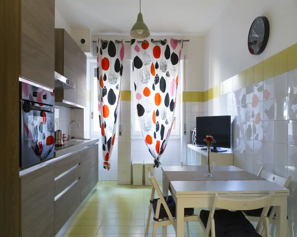 Cocina pequeña luminosa y moderna con muebles efecto madera, horno empotrado, fregadero y una mesa de comedor junto a dos ventanas con cortinas de colores y estampado.