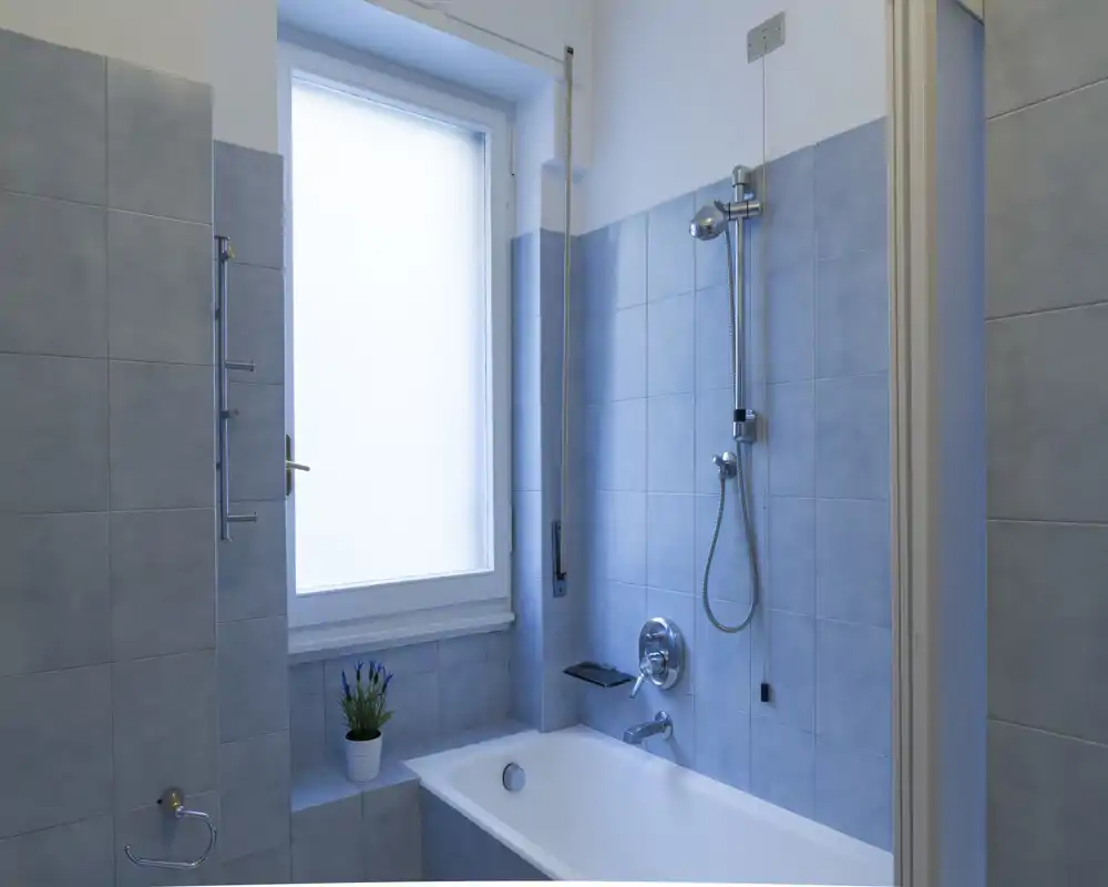 Baño moderno y luminoso con bañera y ducha de pared sobre azulejos azul claro. Ventana translúcida ofrece privacidad dejando entrar luz natural suave.