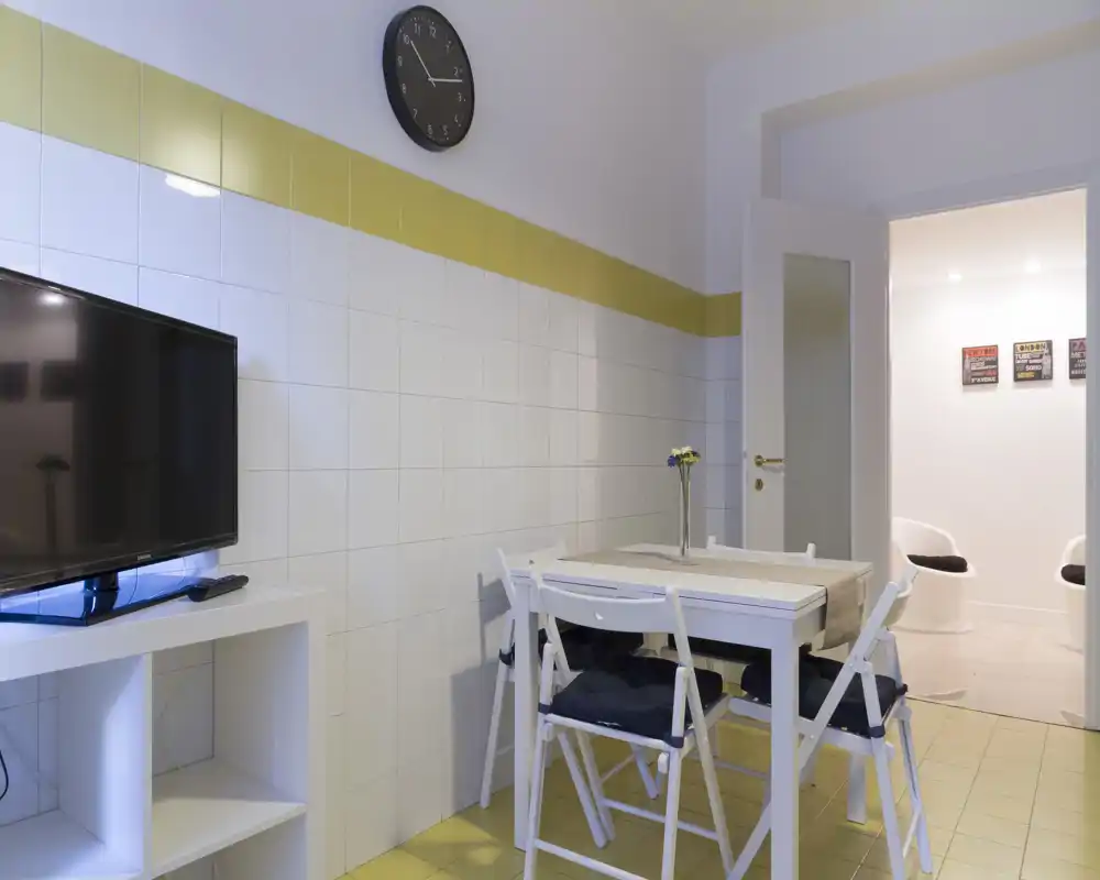 Pequeña zona de comedor con mesa blanca, cuatro sillas plegables con cojines y un pequeño jarrón decorativo; paredes alicatadas y mueble de TV crean un espacio práctico y acogedor.