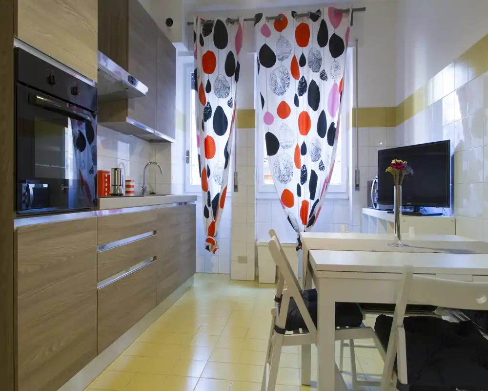 Cocina moderna y luminosa con muebles efecto madera, horno empotrado, fregadero y una pequeña mesa de comedor. Las cortinas estampadas y la televisión crean un espacio acogedor y funcional, ideal para alquileres temporales.