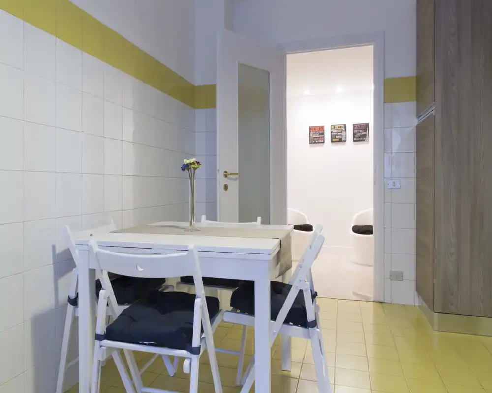 Rincón comedor moderno y luminoso con mesa blanca y sillas plegables sobre suelo de baldosas amarillo claro; un centro de mesa sencillo y líneas limpias crean un área comedor funcional y aireada.