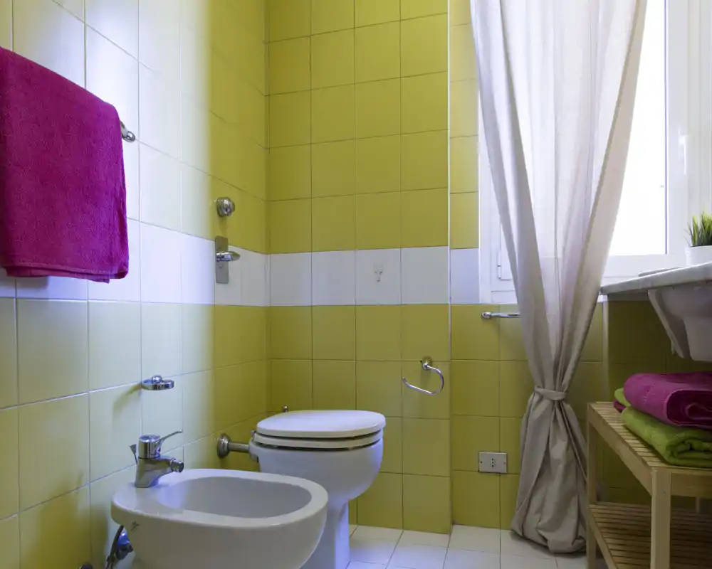 Baño pequeño y luminoso con azulejos amarillos, bidé y WC, lavabo junto a la ventana y toallas dobladas en una estantería de madera.