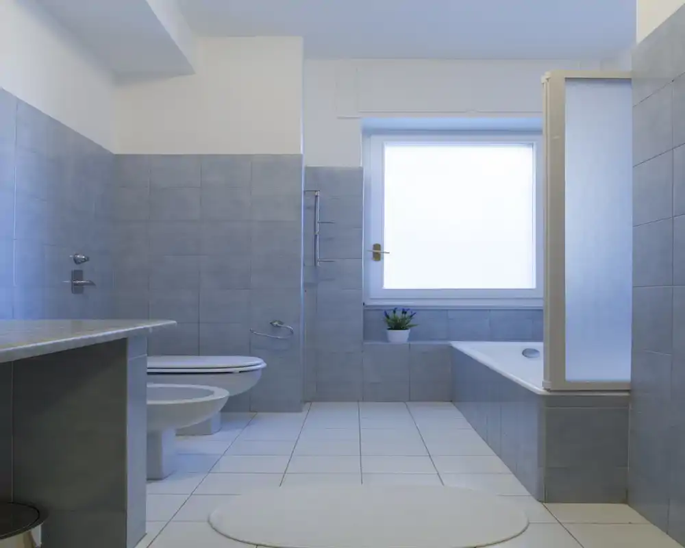 Baño moderno y luminoso con azulejos, bañera con mampara translúcida, inodoro y una gran ventana esmerilada que aporta luz natural suave.