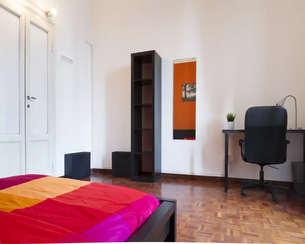 Chambre lumineuse et moderne avec une literie colorée, une étagère simple et un petit bureau — parquet en bois et lignes épurées offrant une impression d'espace minimaliste.