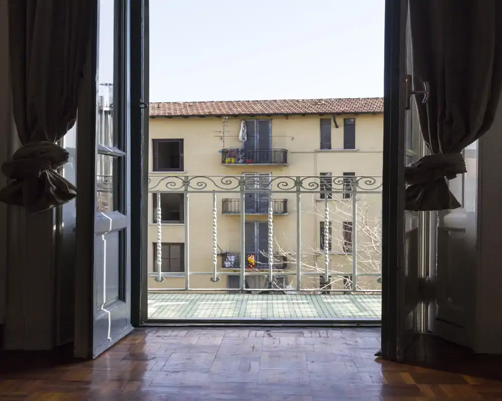 Vue depuis une porte intérieure ouverte sur un petit balcon avec une rambarde en fer décorative et les façades des bâtiments voisins en plein jour.