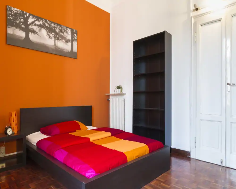 Chambre moderne et lumineuse avec un lit bas double aux couettes rouges, orange et jaune, tête de lit et étagères en bois foncé, et un mur orange accent qui crée une ambiance chaleureuse et contemporaine.