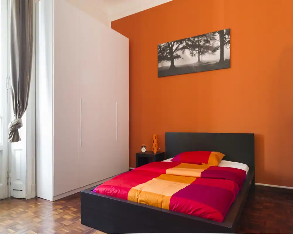 Chambre moderne et lumineuse avec un grand lit bas aux couettes rouges et orange, une armoire blanche et une lumière naturelle provenant d'une haute fenêtre.
