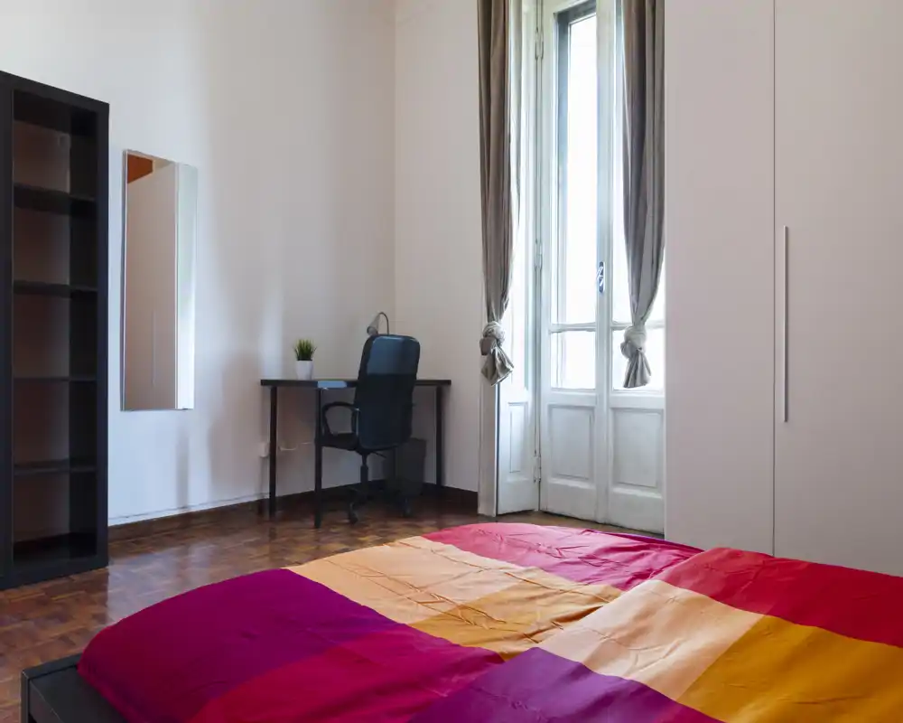 Chambre lumineuse et aérée avec un grand lit au premier plan, parquet, bureau et chaise près d'une grande fenêtre laissant entrer la lumière naturelle. Placard et étagères simples pour le rangement.