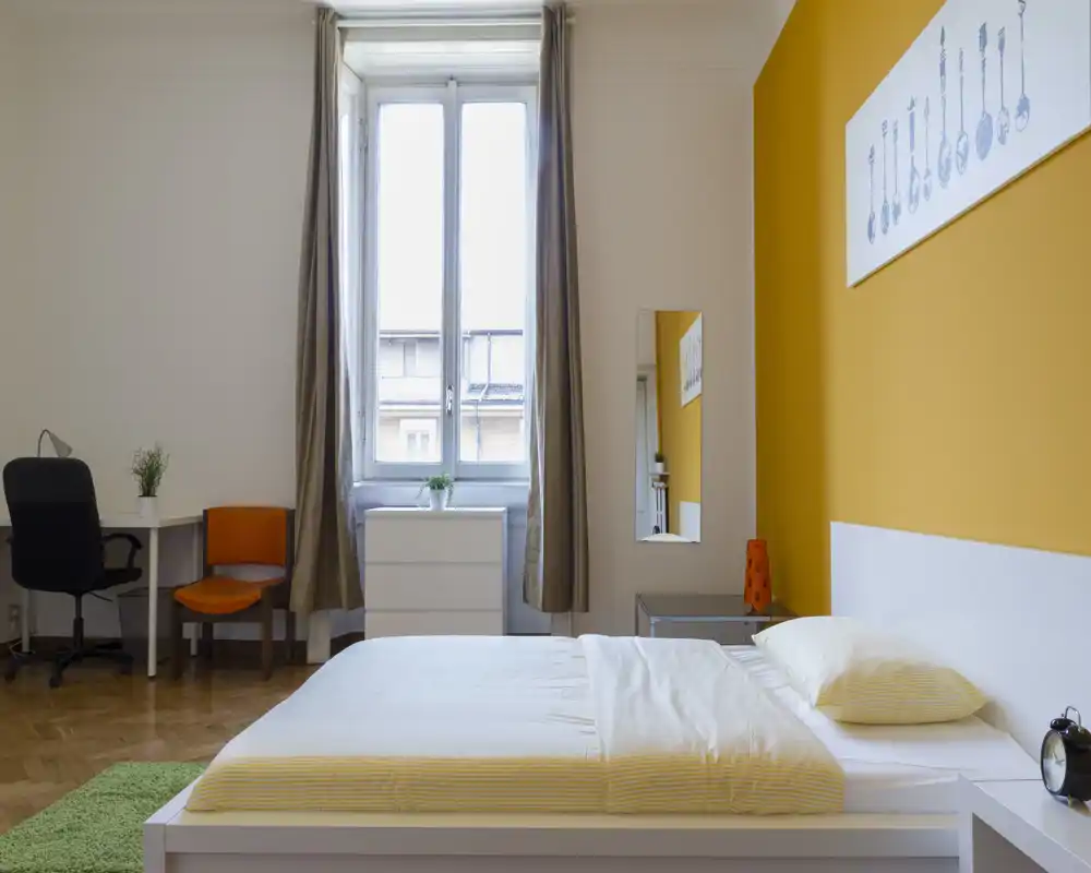 Chambre moderne et lumineuse avec grande fenêtre, lit double confortable en linge clair et mur d'accent jaune; bureau et rangement présents, parfaite pour une annonce locative.