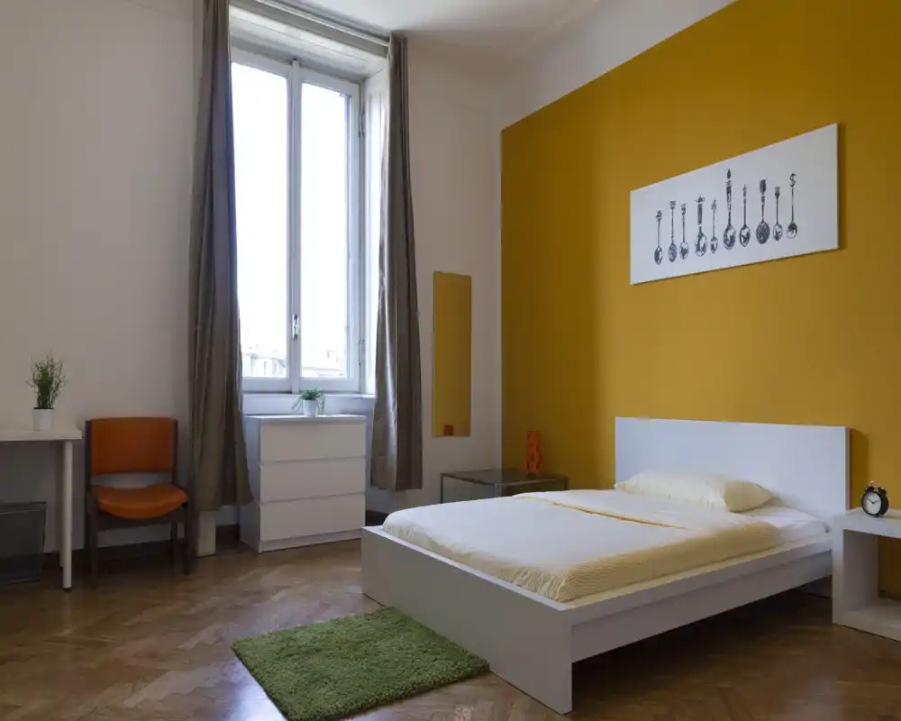 Chambre lumineuse et moderne avec lit plateforme blanc, mur accent jaune et grande fenêtre apportant beaucoup de lumière naturelle. Meubles simples : commode, chaise et table de chevet pour un séjour agréable.