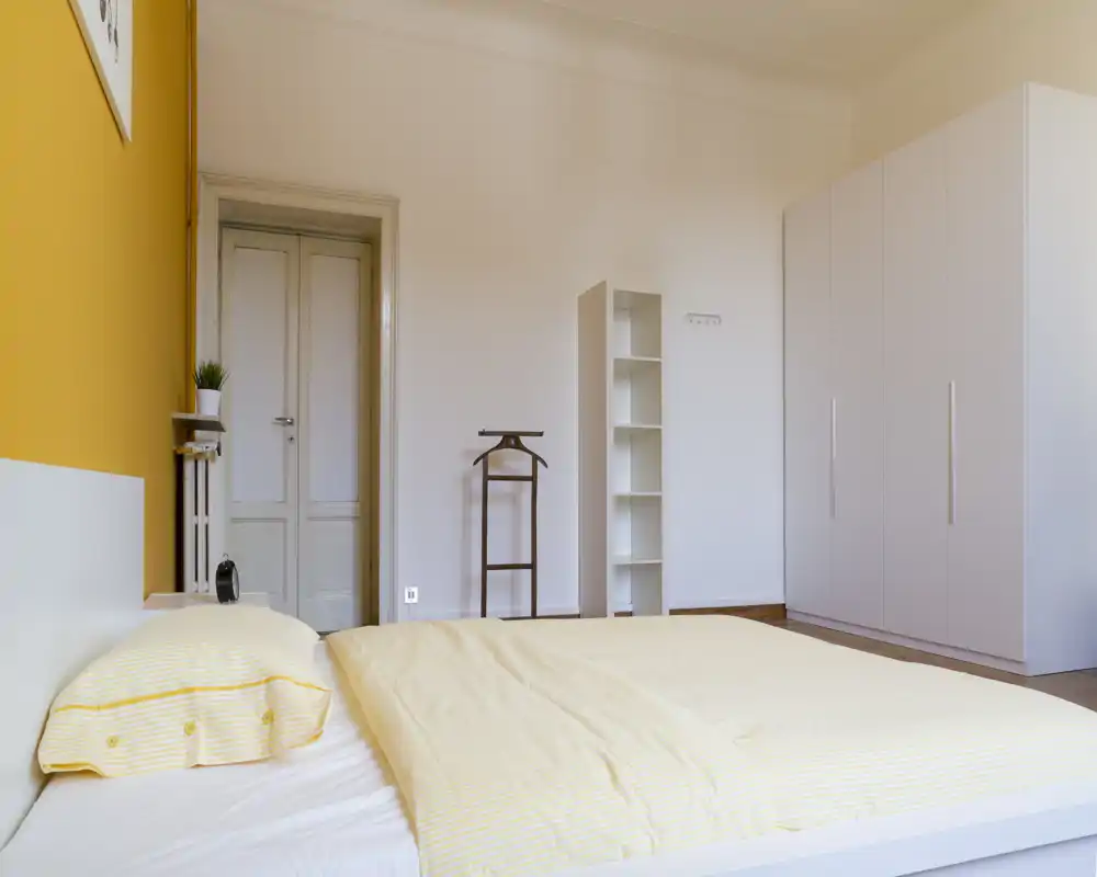 Chambre moderne et lumineuse avec un lit double bas, linge de lit jaune pâle et une grande armoire blanche. L'espace paraît aéré et chaleureux grâce à une décoration épurée et un mur d'accent chaud.