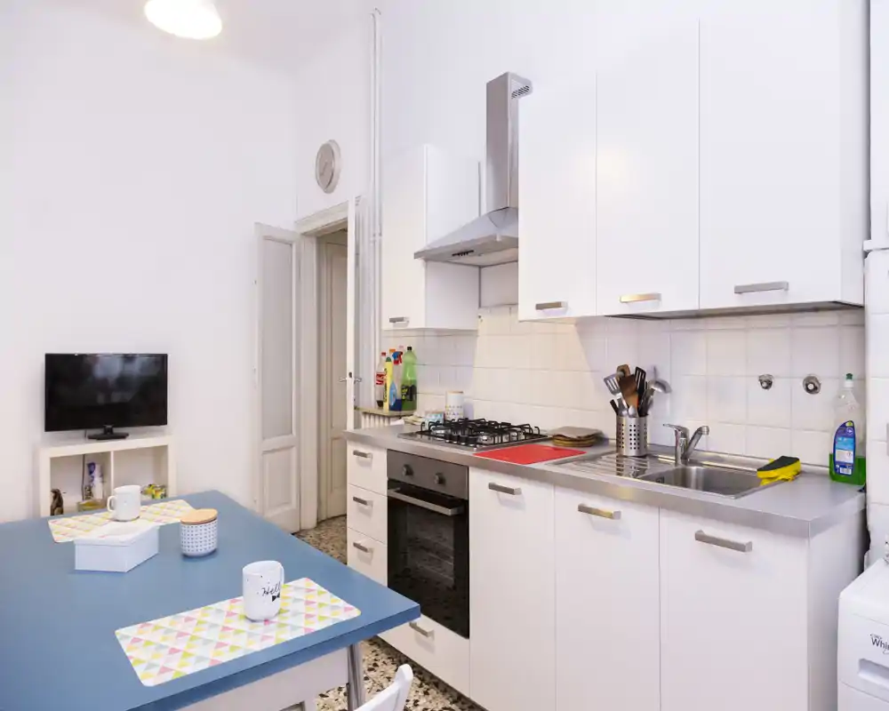 Cuisine compacte et moderne avec meubles blancs, évier en inox, plaque à gaz et table équipée de sets et tasses — lumineuse et bien organisée pour un usage quotidien.