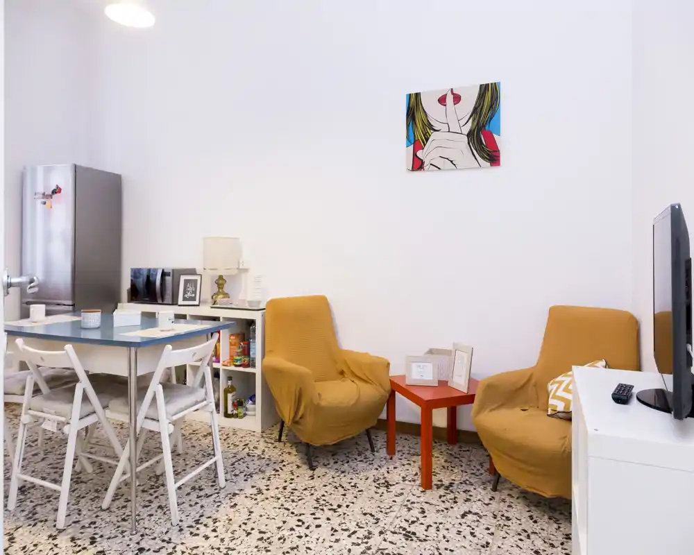 Coin salon chaleureux avec deux fauteuils moutarde autour d'une petite table basse rouge, une télévision et une table à manger; murs blancs lumineux et un tableau coloré apportent une atmosphère accueillante.