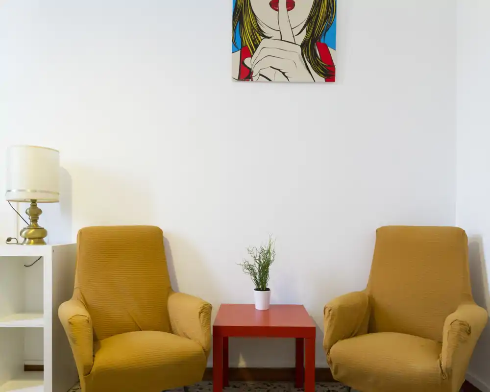 Espace de vie lumineux et minimaliste avec deux fauteuils moutarde face à une petite table d'appoint rouge et une lampe, idéal pour un coin lecture chaleureux.