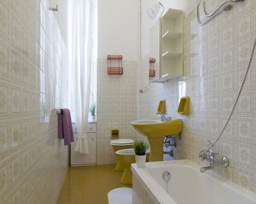 Salle de bain rétro lumineuse avec carrelage à motifs, baignoire avec douchette, lavabo, bidet et WC jaune assortis, et une grande fenêtre laissant entrer la lumière naturelle.