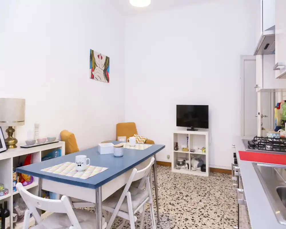 Espace de vie lumineux et bien rangé avec table à manger et kitchenette compacte. On y voit une table bleue, des chaises pliantes, une petite TV sur étagère blanche et des assises avec coussins — parfait pour un appartement cosy.