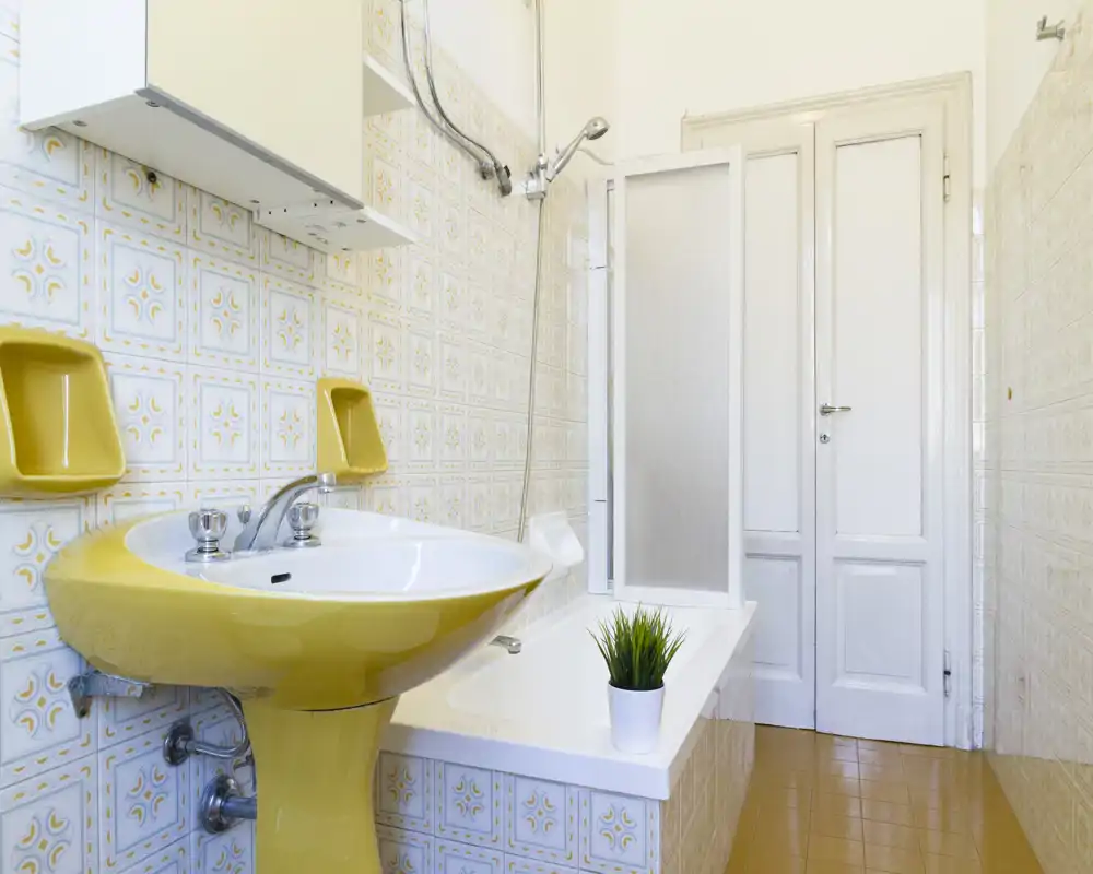 Salle de bain lumineuse au style rétro avec lavabo jaune sur colonne, baignoire avec paroi de douche et carrelage à motifs. Agencement propre et bien éclairé.