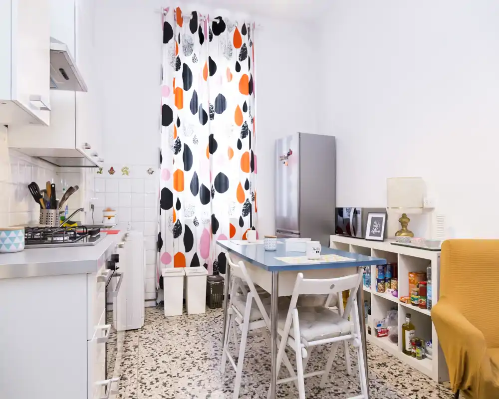 Cuisine compacte et lumineuse avec cuisinière à gaz, réfrigérateur, table à manger et rideaux colorés ; espace soigné et accueillant, idéal pour un petit appartement.
