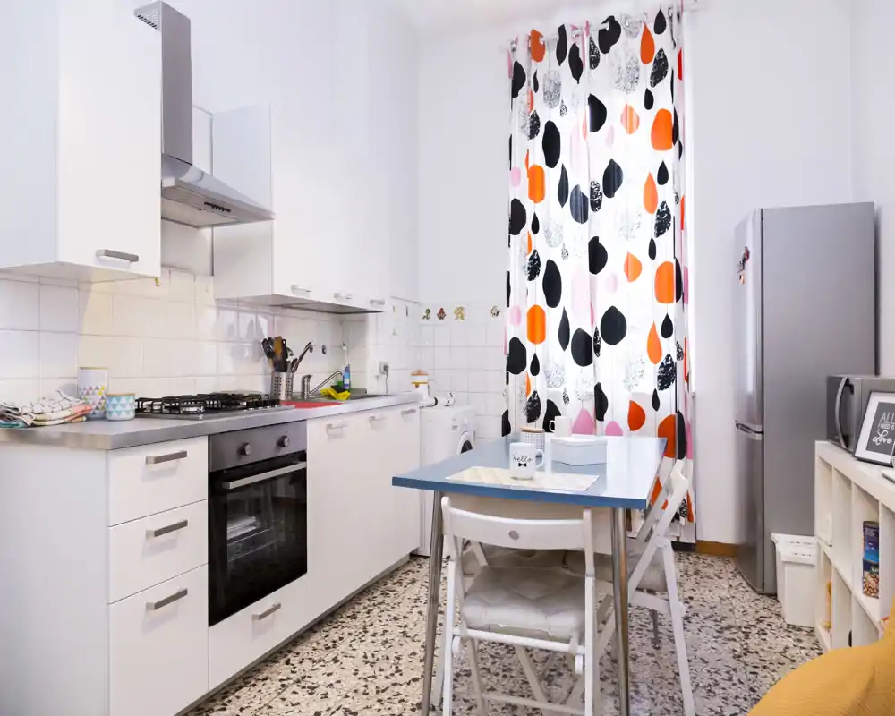 Cuisine compacte et lumineuse avec placards blancs, cuisinière à gaz, réfrigérateur et une petite table à manger devant un rideau coloré à motifs.