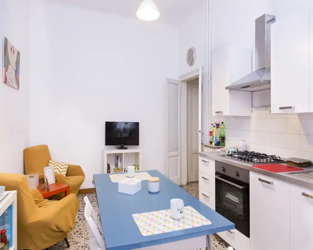 Espace de vie lumineux en open-space avec coin cuisine. Table bleue, deux fauteuils confortables et kitchenette moderne avec plaque à gaz — idéal pour un petit appartement.