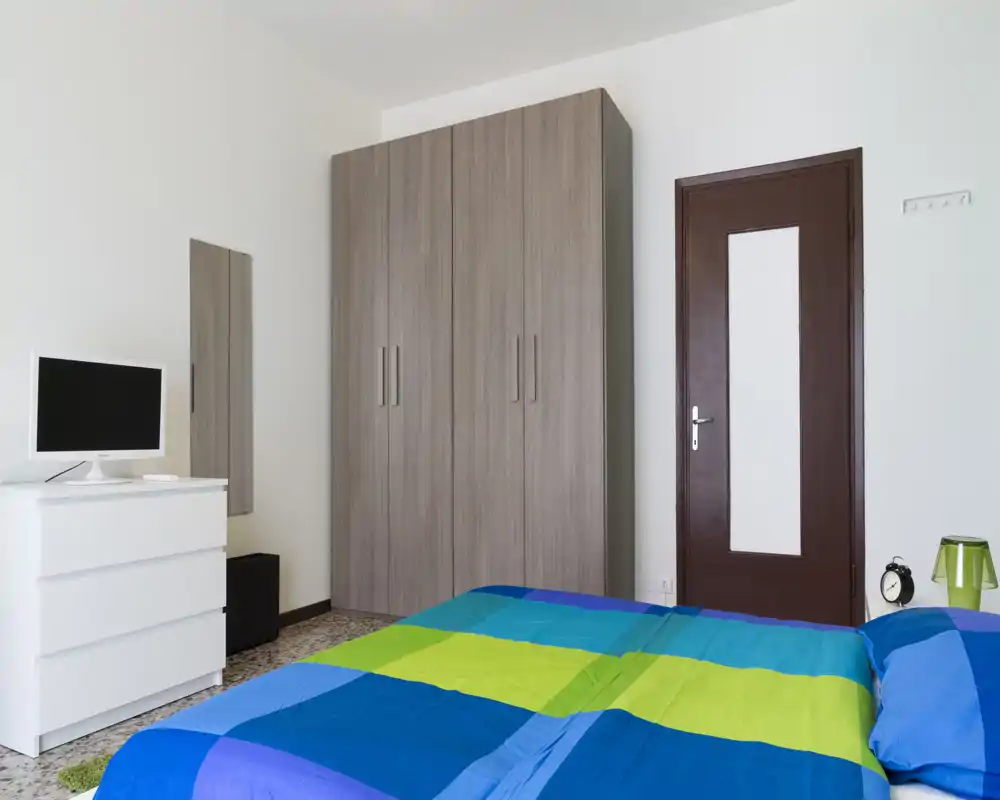 Bright, modern bedroom with a large wardrobe, white chest of drawers, and a colorful blue-green bedspread. Clean, minimal styling and good natural light, suitable for rental listings.