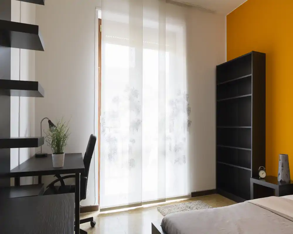 Bright, modern bedroom with a large window covered by sheer panels, a comfortable bed at the right foreground, desk and shelving for work, and a warm orange accent wall.