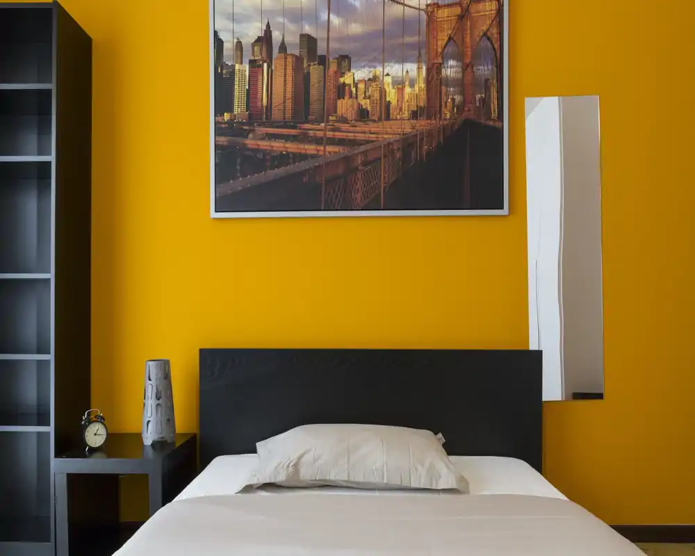 Bright, modern bedroom with a neatly made bed, dark headboard and warm orange accent wall featuring a cityscape artwork.
