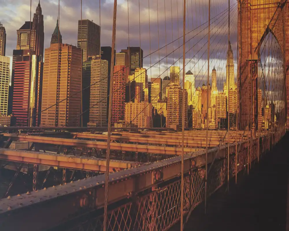 A dramatic outdoor cityscape showing a bridge with suspension cables in the foreground and a skyline of tall buildings bathed in warm light.