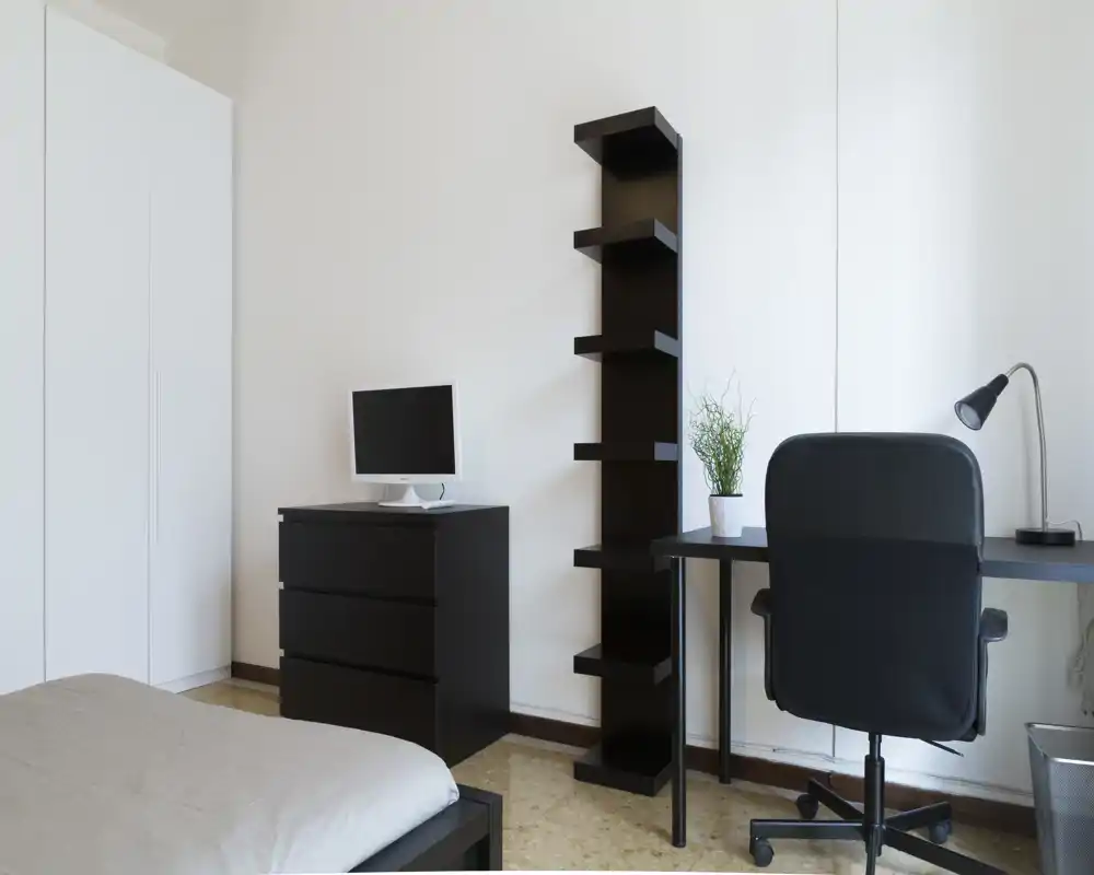 Modern bedroom with a visible bed corner, black storage furniture, and a compact desk with office chair — bright, minimalist and functional.